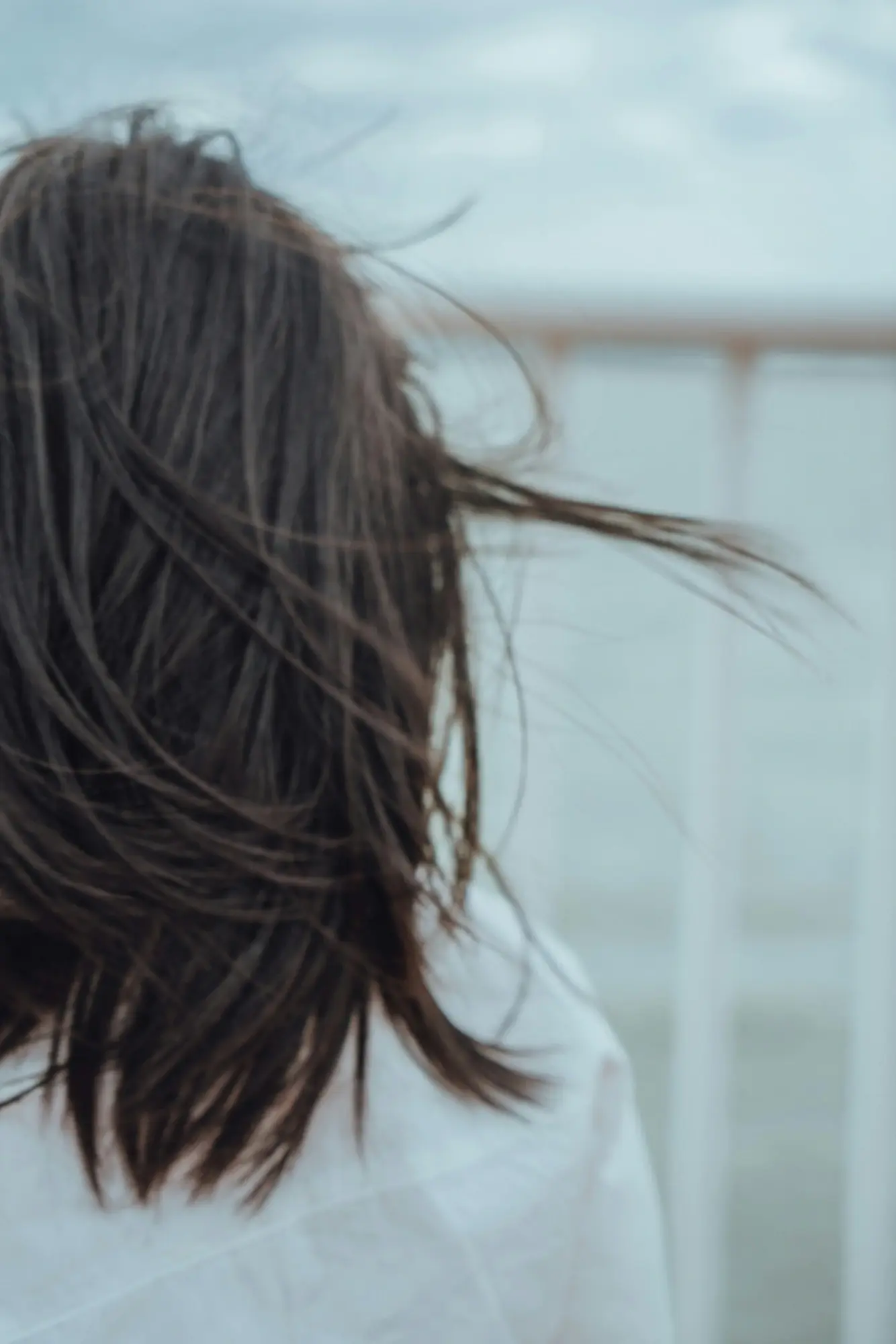 A woman with long hair stands by the ocean, gazing at the waves under a clear blue sky.